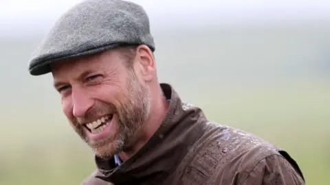 PA Prince William wearing a brown wax coat and gray wool cap. He has a beard and is smiling at the camera