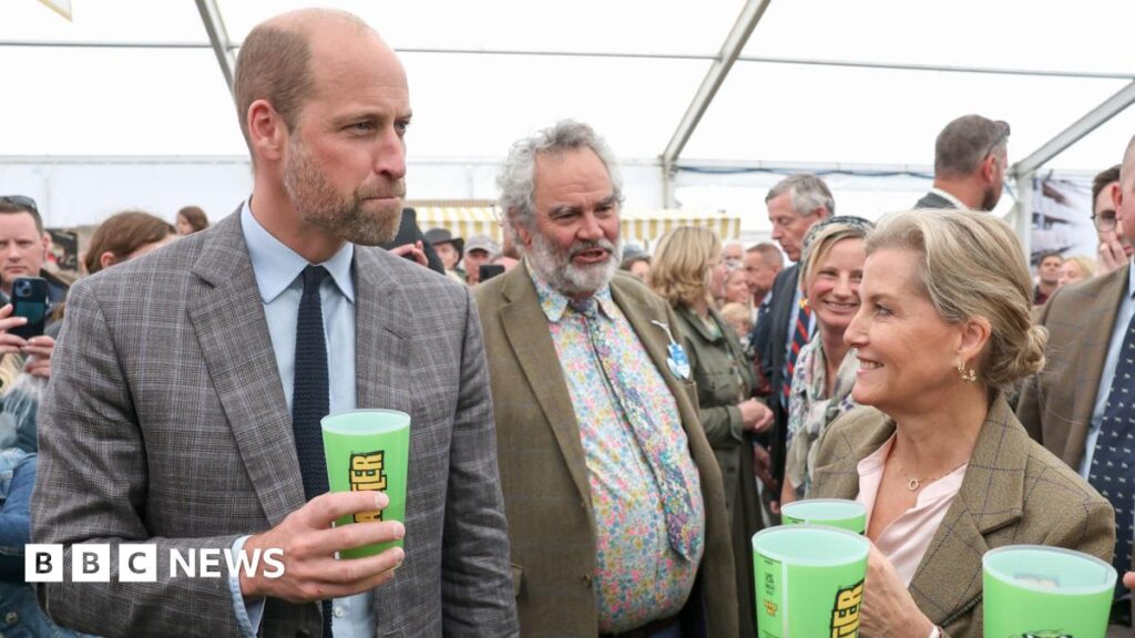 The Prince of Wales at the Royal Cornwall Show with his aunt Sophie, Duchess of Edinburgh. They are standing inside a tent and is surrounded by visitors to the show. They are both trying Rattler cider from a green plastic cup.