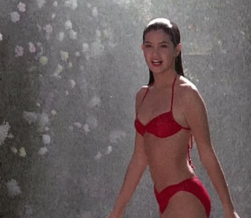Phoebe Cates in a red bikini poses confidently under sprinkling water, exuding a glamorous and relaxed vibe