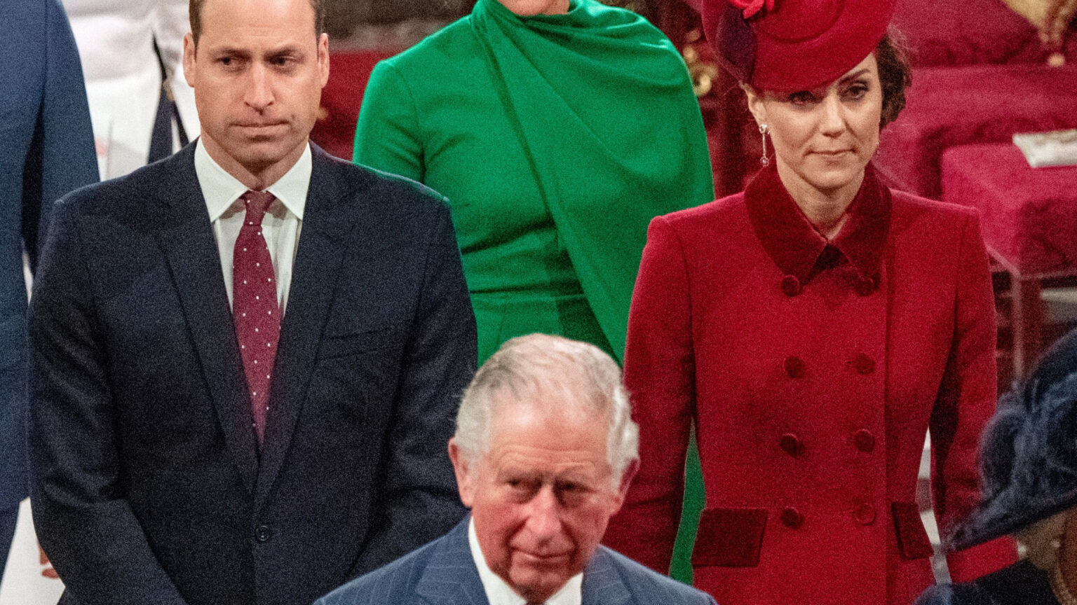 Prince William is wearing a dark suit with a red tie, while Kate Middleton wears a red coat dress and matching hat, and King Charles walks ahead of them in a striped blue suit