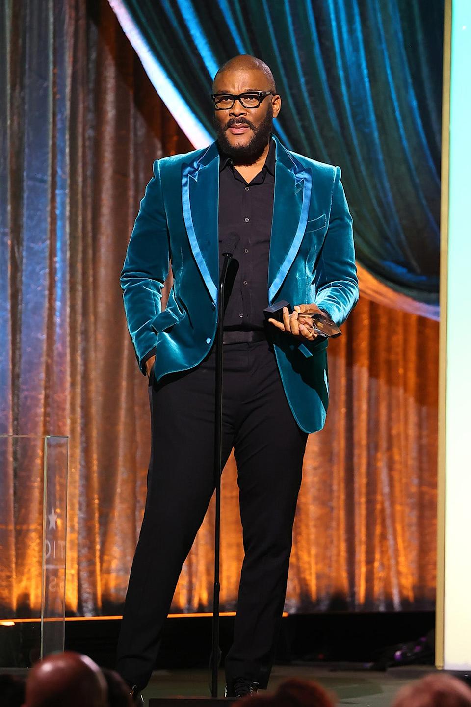 Tyler Perry wearing a bright blue velvet coat and a dark suit speaking on stage.