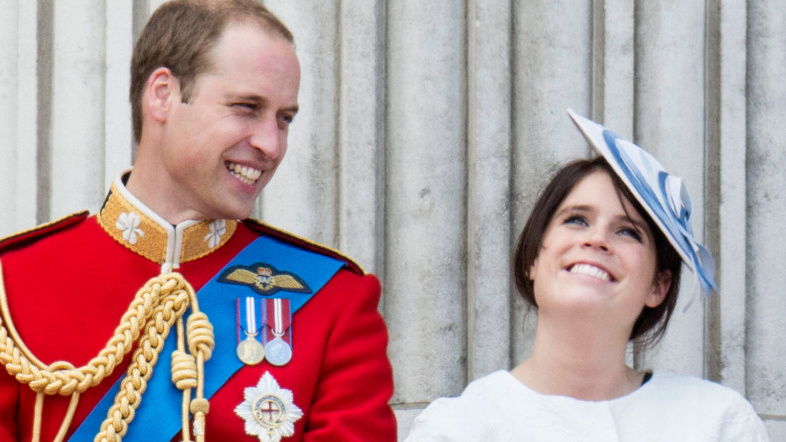 Princess Eugenie’s Unexpected Alliance With Prince William Prince William laughs while wearing red military uniform as he stands next to Princess Eugenie, who is wearing a white dress and a blue and white hat on the Buckingham Palace balcony