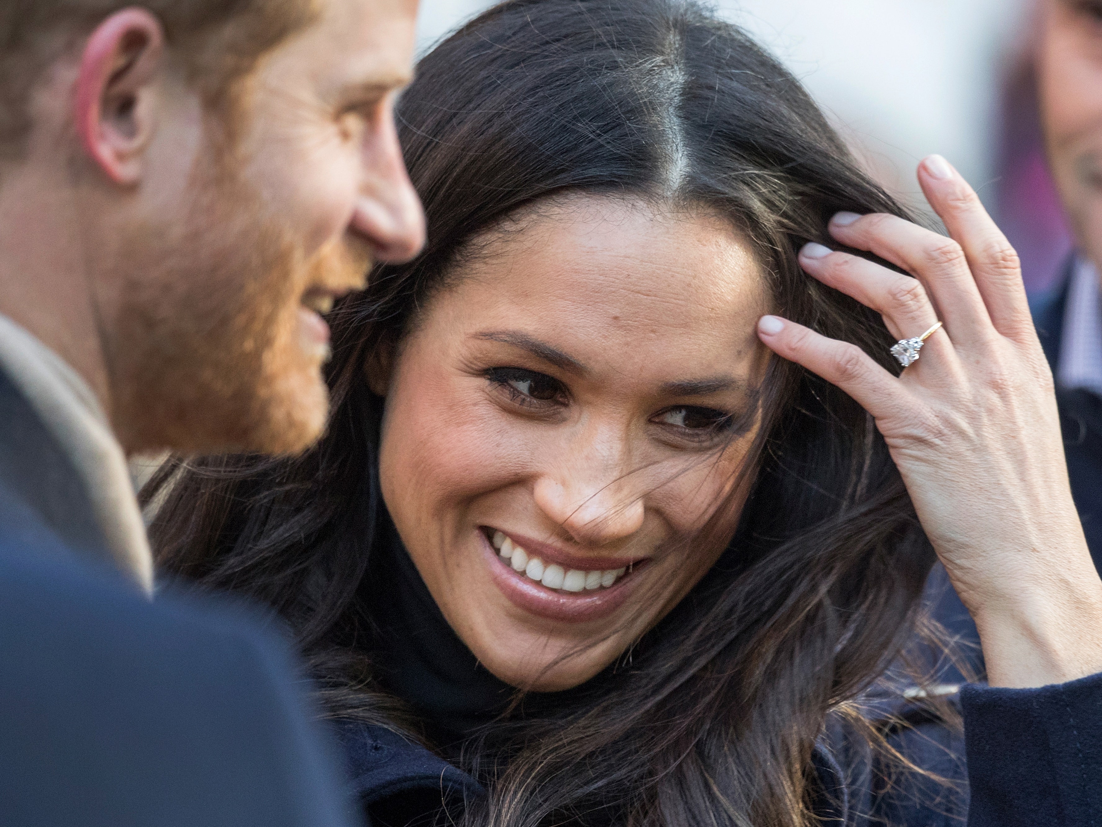 Prince Harry, with a beard, looks towards Meghan Markle, who is smiling and has her hand near her hair, displaying her engagement ring.