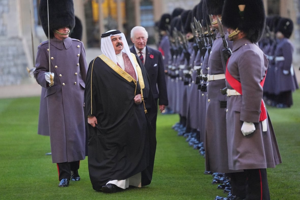 Two people walk on grass in front of a line of people wearing military uniforms.