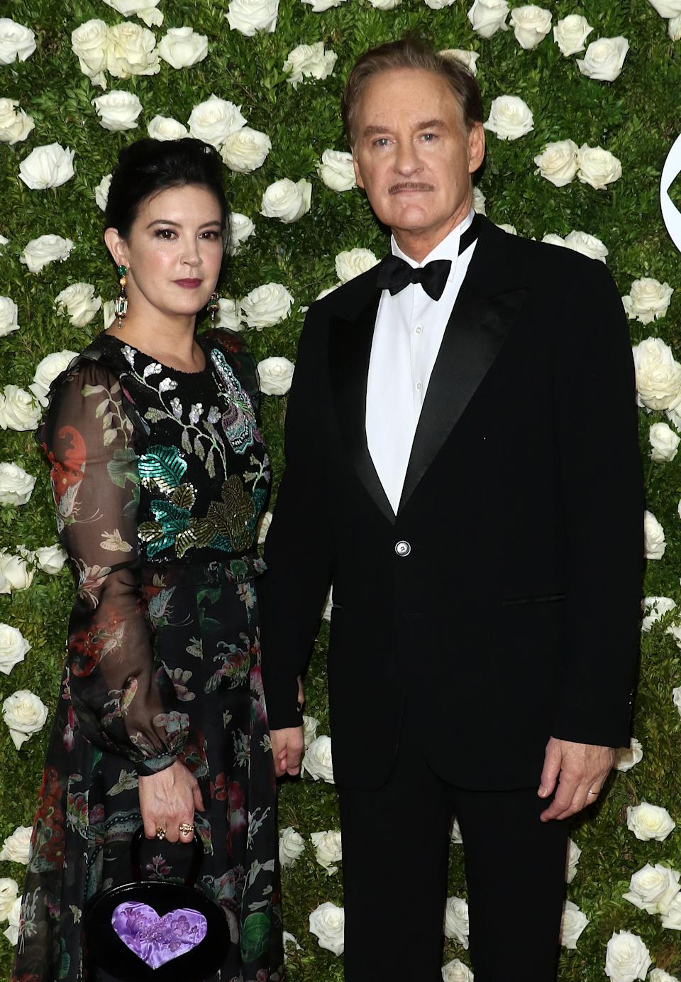 Phoebe Cates and Kevin Kline stand on a floral backdrop. The woman wears a floral dress, and the man is in a classic tuxedo