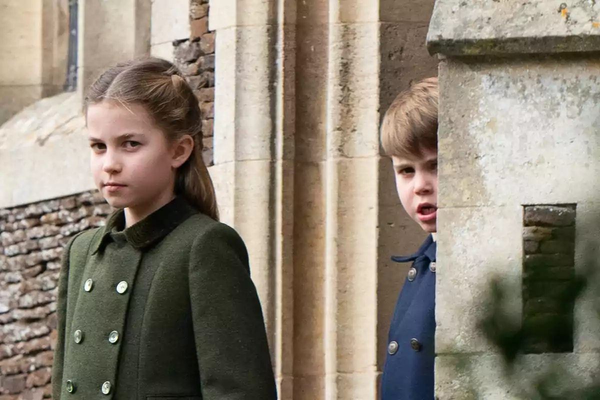 Princess Charlotte and Prince Louis in stylish coats are standing next to a stone wall.