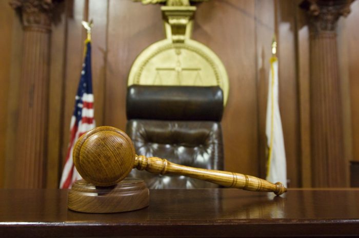 A wooden judge's gavel rests on a wooden desk in a courtroom, with an empty judge's chair and flags in the background.