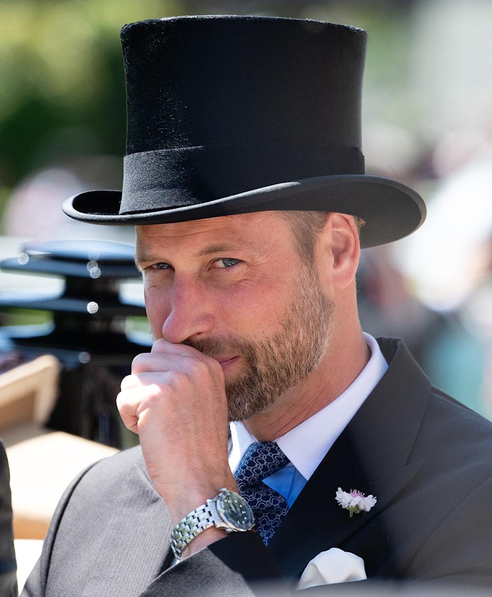 Prince William with his fist to his face as he smirks with a dark suit and a black top hat.