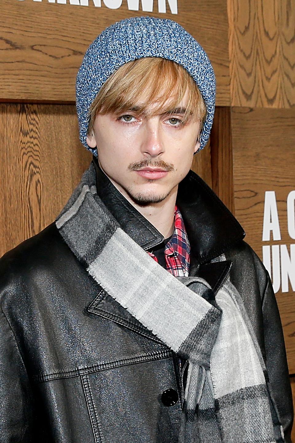 Timothée Chalamet in a beanie and plaid shirt, wearing a leather jacket with a checked scarf, at a formal event