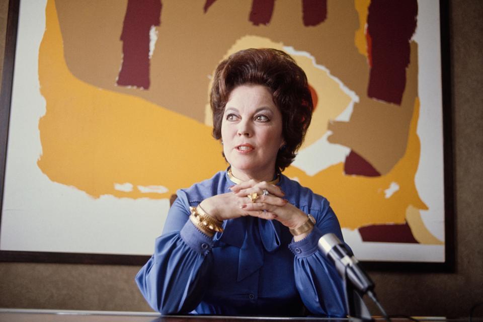 Adult Shirley Temple seated at a desk with a microphone, wearing a stylish blouse and bracelets, with abstract art in the background