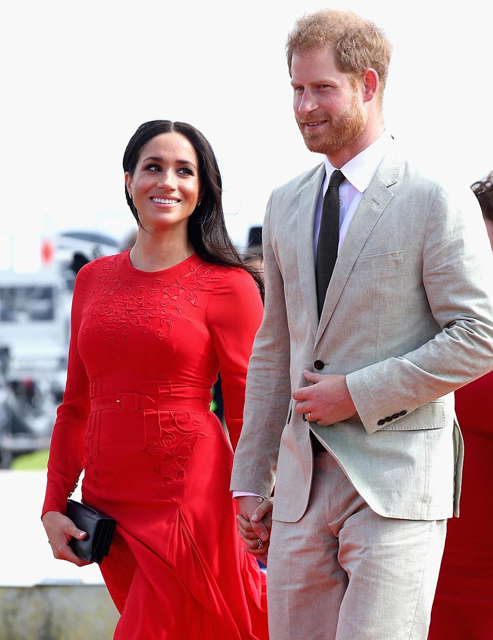 Meghan Markle wearing a red dress smiling at Prince Harry wearing a grey suit and dark tie.