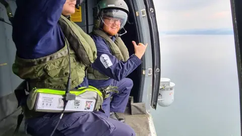 Aran Woolford Aran Woolford is crouching and smiling inches away from a doorway of an aircraft in flight. He is wearing a flying suit and helmet and is next to a colleague. The sea can be seen below. 