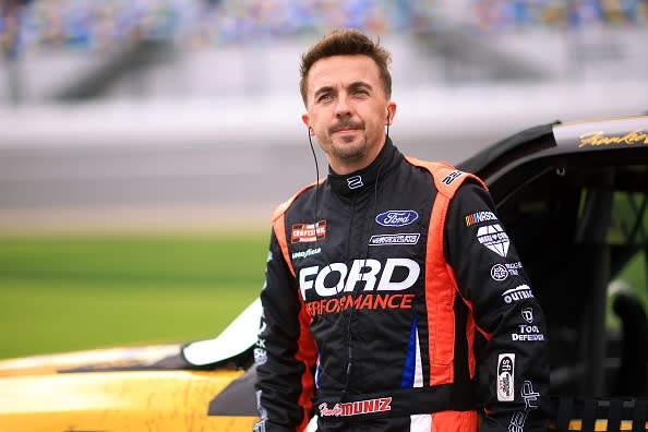 Frankie Muniz posing beside a race car, wearing a Ford Performance racing suit with sponsor logos, looking confident