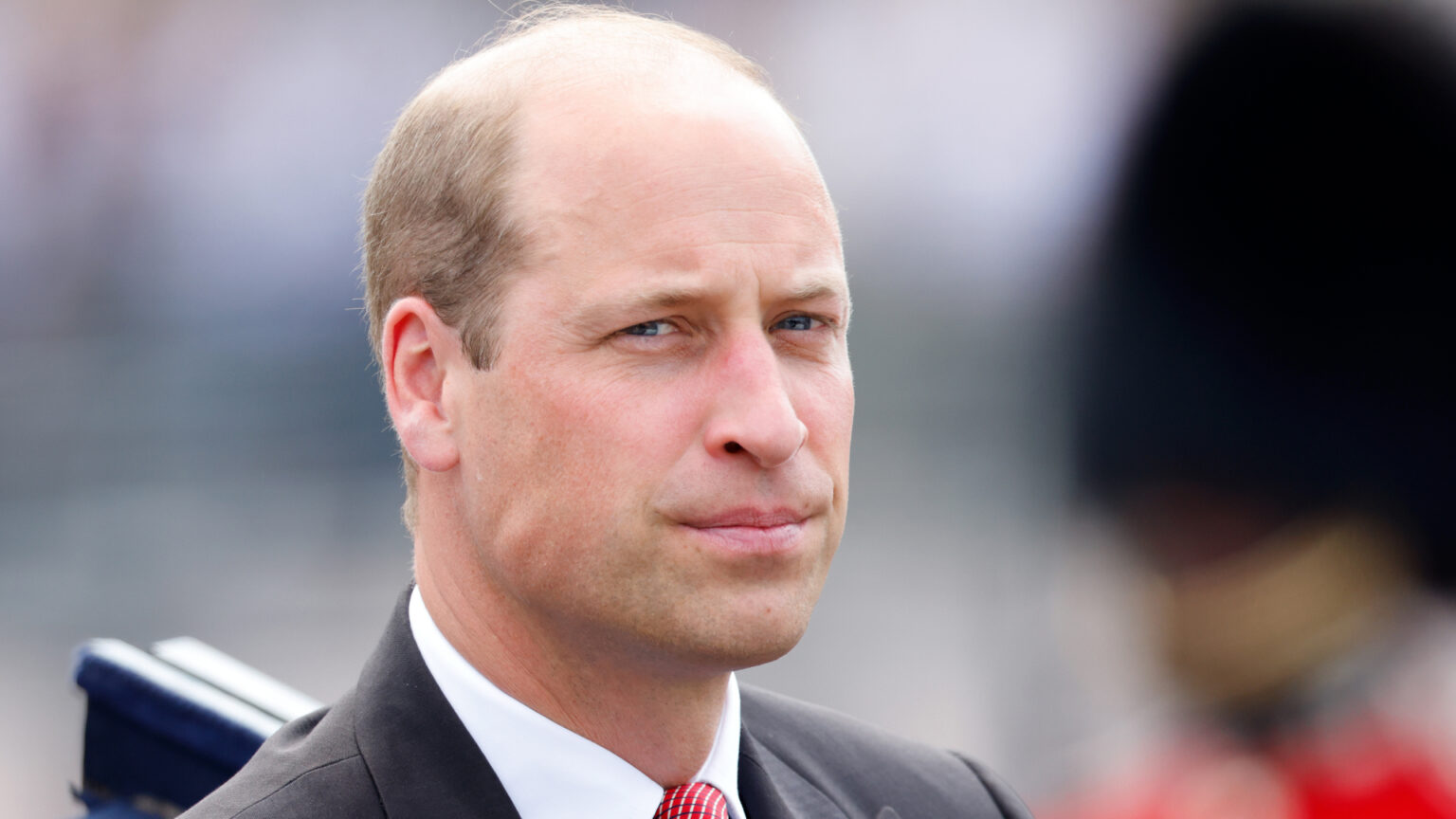Prince William wears a black suit with a red tie and looks serious