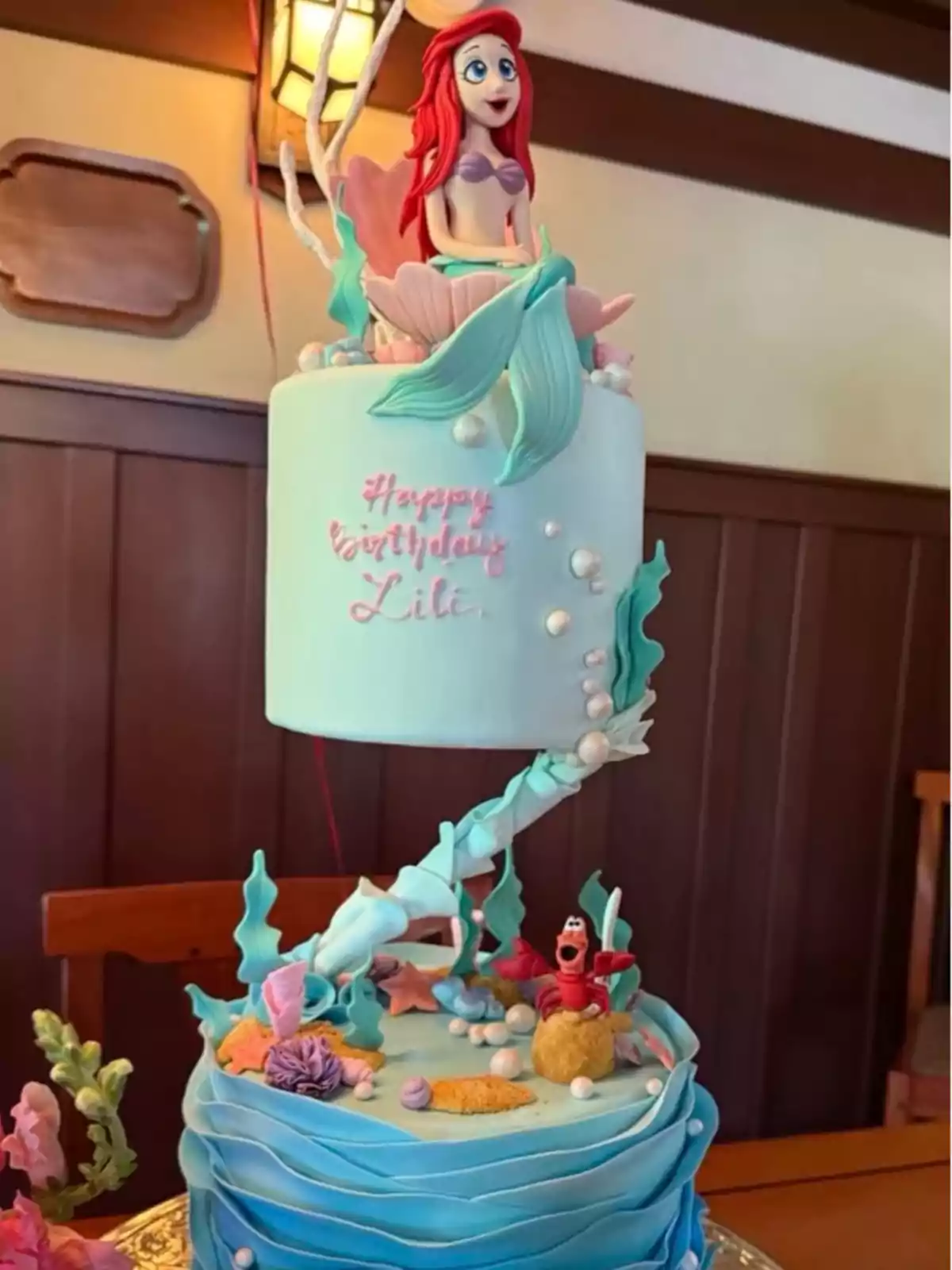 Two-tier cake decorated with a Little Mermaid theme, featuring an Ariel figurine on top and marine details like corals, pearls, and a red crab at the base, along with a birthday message for Lili. Two-tier cake decorated with a Little Mermaid theme, featuring an Ariel figurine on top and marine details like corals, pearls, and a red crab at the base, along with a birthday message for Lili.