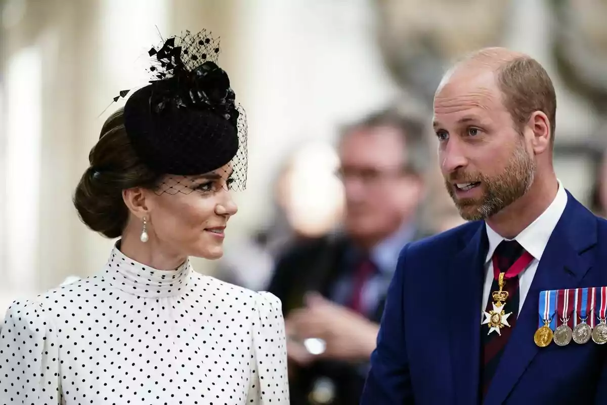 A woman in a white polka-dot dress and a black hat is talking to a man in a blue suit with medals and decorations at a formal event.