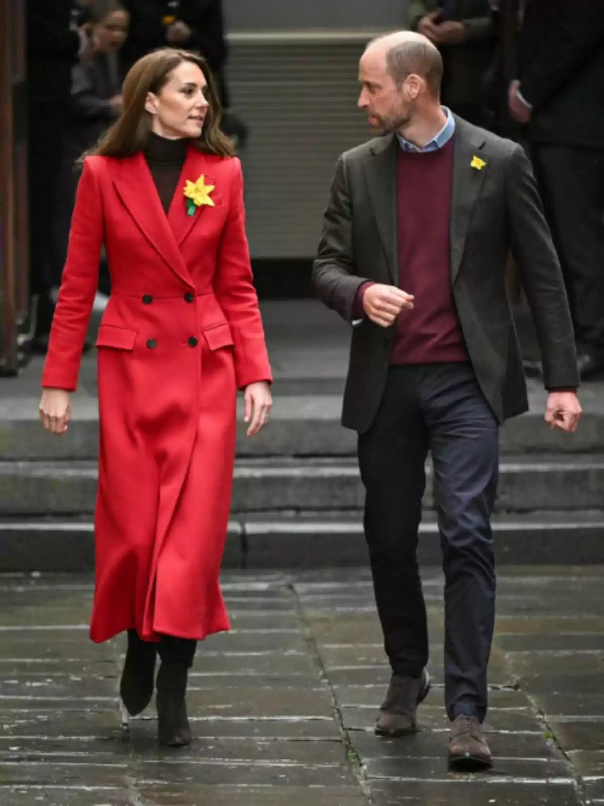 Kate Middleton and Prince William walking together on a street, one of them wearing a red coat and the other a burgundy sweater with a dark jacket, both have yellow flowers on their lapel.