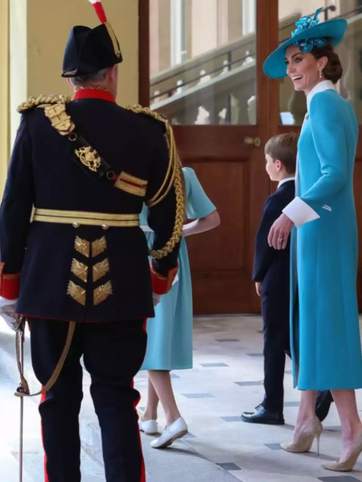 An elegantly dressed woman in blue with a matching hat smiles as she talks with a man in a formal uniform and two children in an official-looking building.