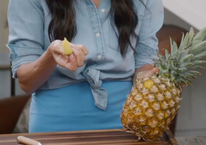 Woman demonstrating a kitchen hack by peeling a pineapple while wearing a blue shirt and apron in a home kitchen. Woman demonstrating a kitchen hack by peeling a pineapple while wearing a blue shirt and apron in a home kitchen.