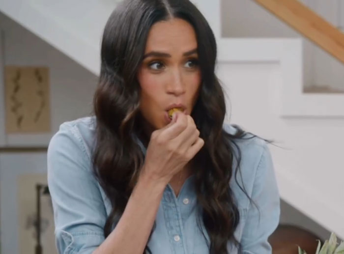 Meghan Markle eating a snack indoors, wearing a blue shirt, sharing a kitchen hack in a casual home setting. Meghan Markle eating a snack indoors, wearing a blue shirt, sharing a kitchen hack in a casual home setting.