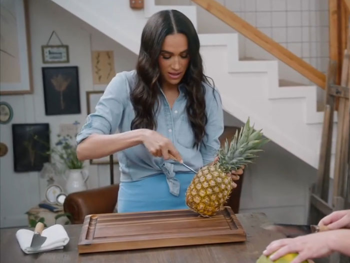 Meghan Markle demonstrating a kitchen hack by cutting a pineapple on a wooden board in a home setting. Meghan Markle demonstrating a kitchen hack by cutting a pineapple on a wooden board in a home setting.
