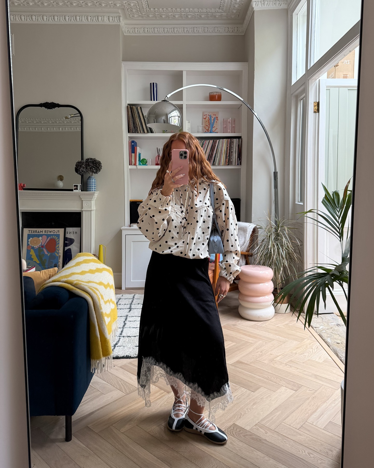Influencer takes a mirror selfie instead wearing a white polkadot blouse with a black lace-trim skirt and strappy sneakerinas.
