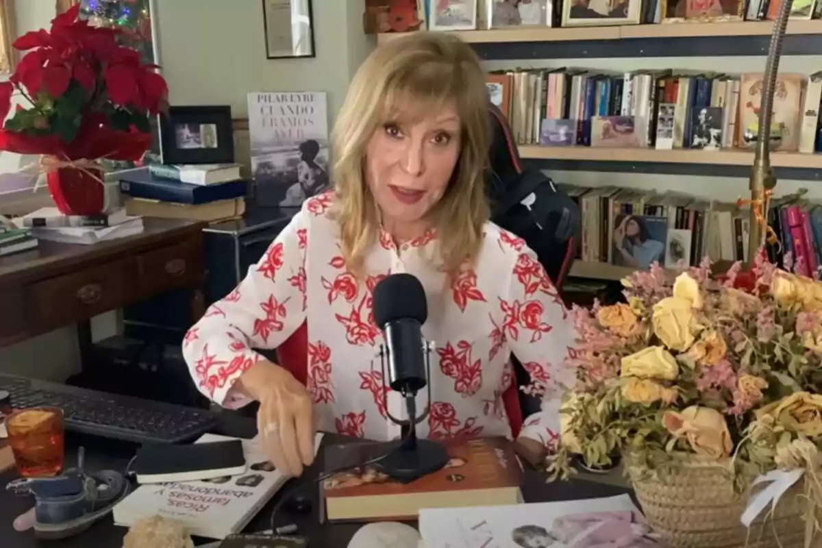 A blonde-haired woman in a white blouse with red flowers sits in front of a microphone in a room filled with books and decorations, including a red poinsettia plant and a bouquet of dried flowers. A blonde-haired woman in a white blouse with red flowers sits in front of a microphone in a room filled with books and decorations, including a red poinsettia plant and a bouquet of dried flowers.