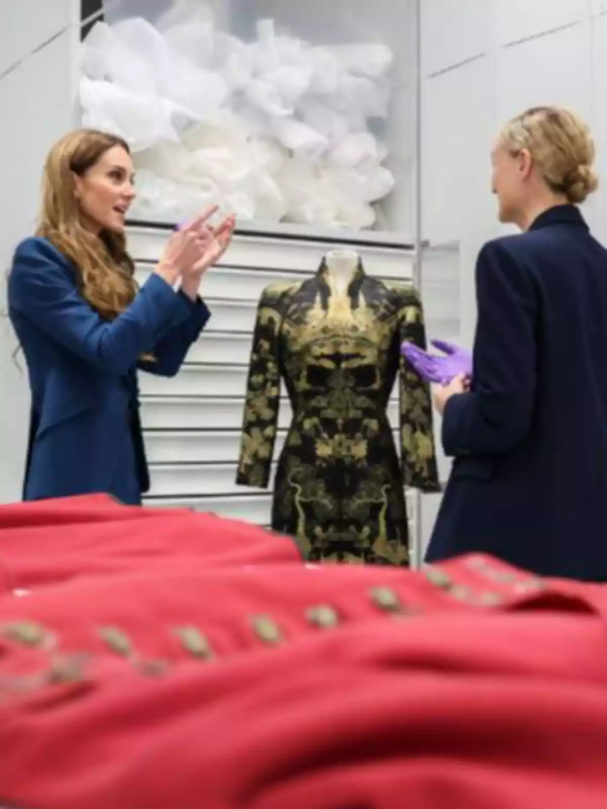 Kate Middleton and another woman are talking in front of a mannequin with a black and gold dress in a room where there are several red garments on a table. Kate Middleton and another woman are talking in front of a mannequin with a black and gold dress in a room where there are several red garments on a table.