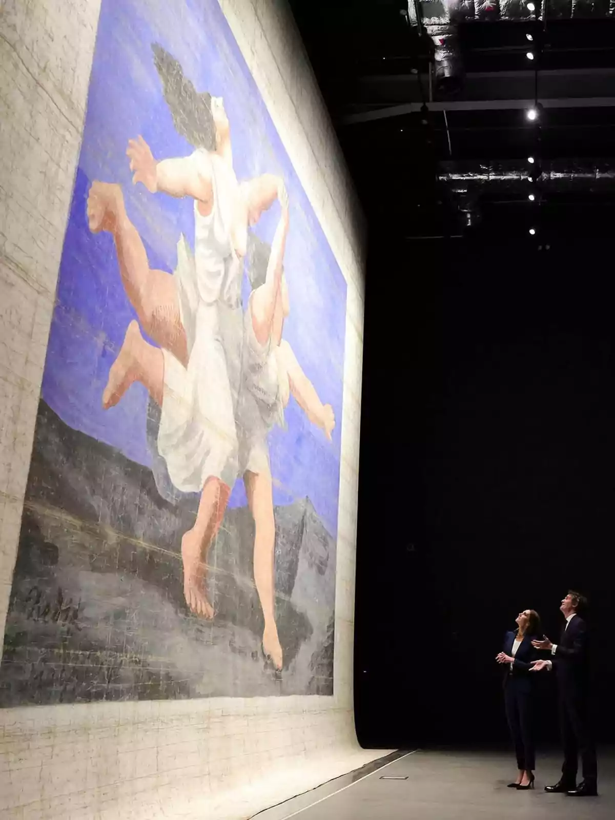 Kate Middleton and a man observe a large painted backdrop featuring the image of two female figures in motion against a blue, mountainous background inside a dark, spacious area. Kate Middleton and a man observe a large painted backdrop featuring the image of two female figures in motion against a blue, mountainous background inside a dark, spacious area.