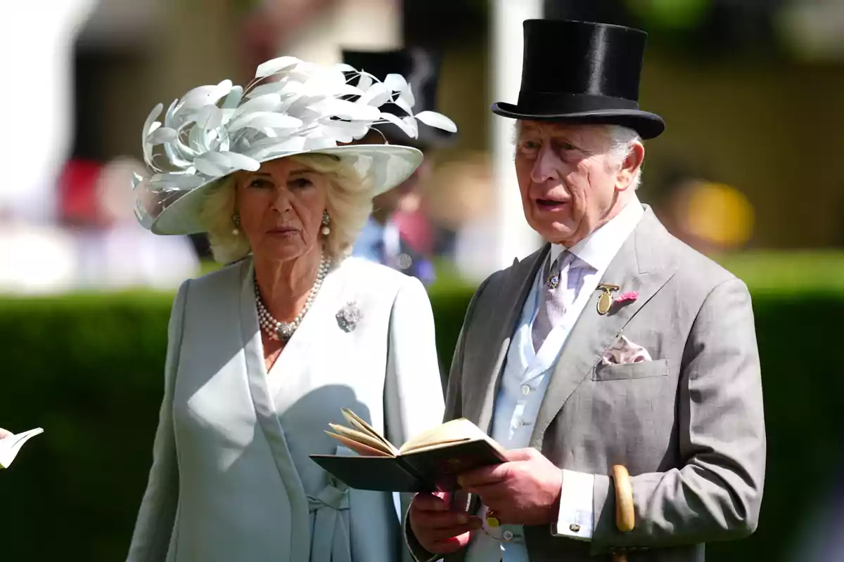Queen Camilla and Charles III, elegantly dressed, attend an outdoor event; the woman wears a decorative white hat and the man a black top hat and holds a book.