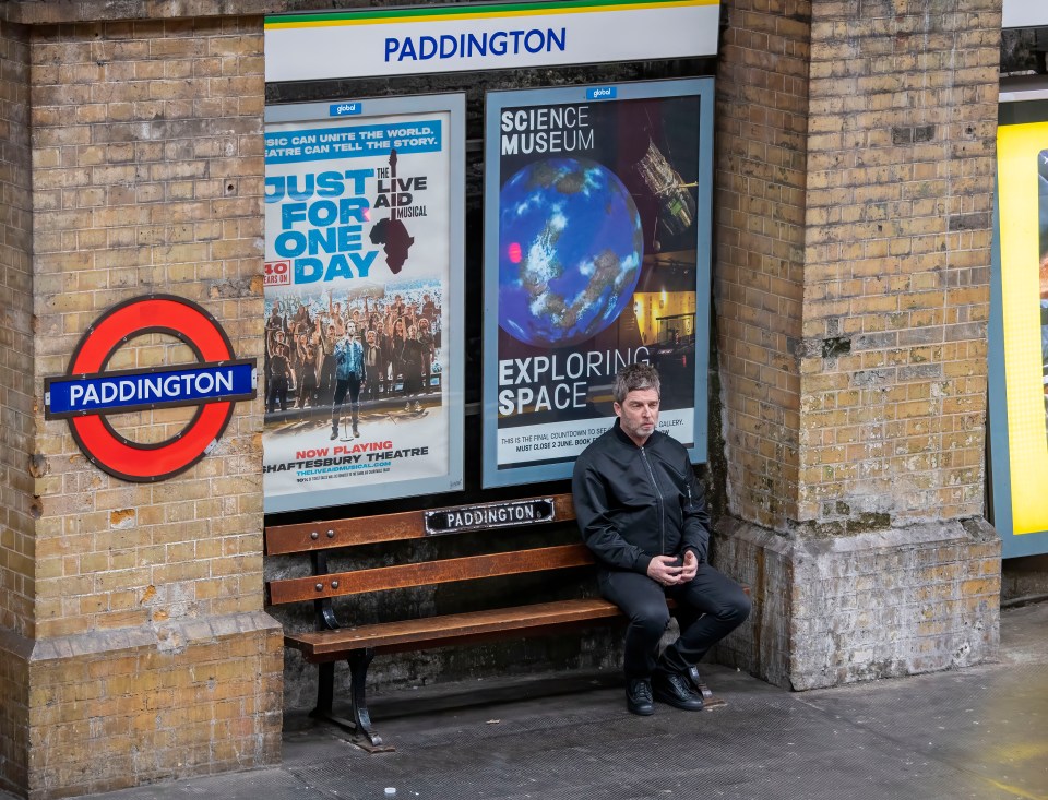 Noel Gallagher sitting on a bench at Paddington Station.