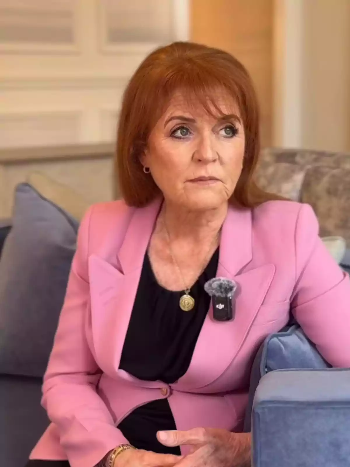 Red-haired woman sitting on a sofa, wearing a pink blazer and a black blouse, looking serious and wearing a lapel microphone.