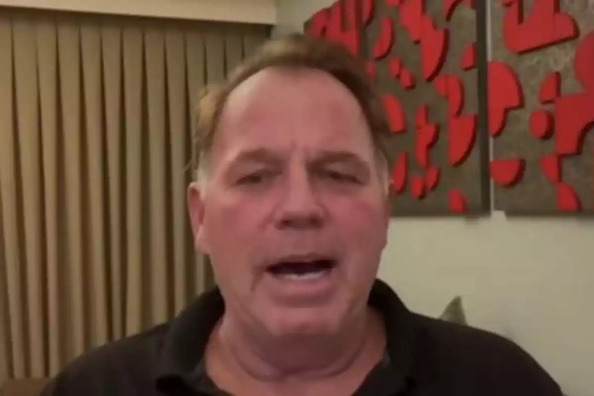 Thomas Markle Jr. in a dark jersey is speaking in front of a beige curtain and a wall decorated with red geometric shapes.