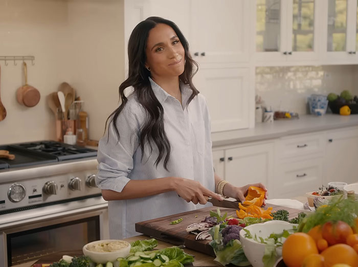 Meghan Markle in kitchen sharing cooking hack, preparing vegetables on wooden cutting board with fresh produce nearby Meghan Markle in kitchen sharing cooking hack, preparing vegetables on wooden cutting board with fresh produce nearby