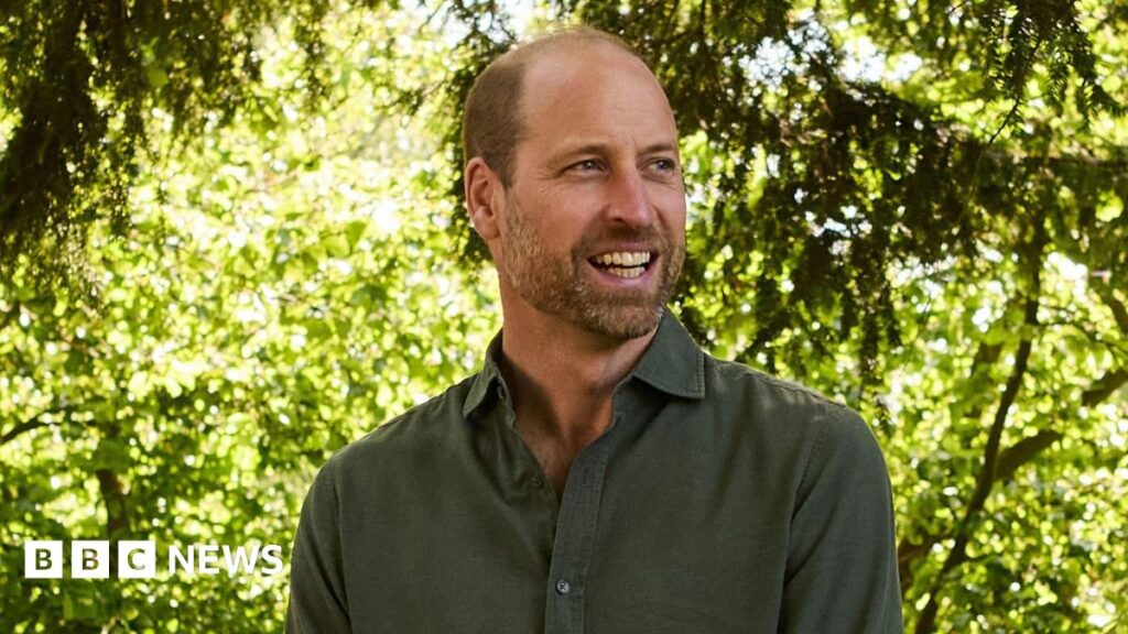 Prince William is wearing a dark green shirt. He is looking away from the camera and smiling. The background is out of focus but has greenery and trees in the background.