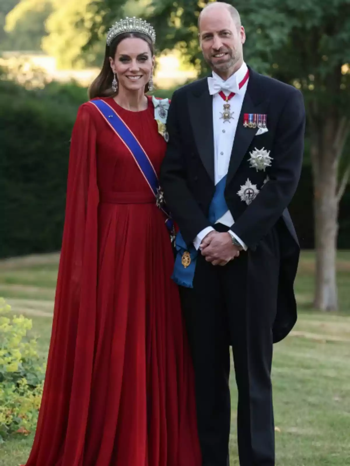 A woman in a long red dress and tiara stands next to a man in a tuxedo with medals as they pose outdoors in a garden. A woman in a long red dress and tiara stands next to a man in a tuxedo with medals as they pose outdoors in a garden.