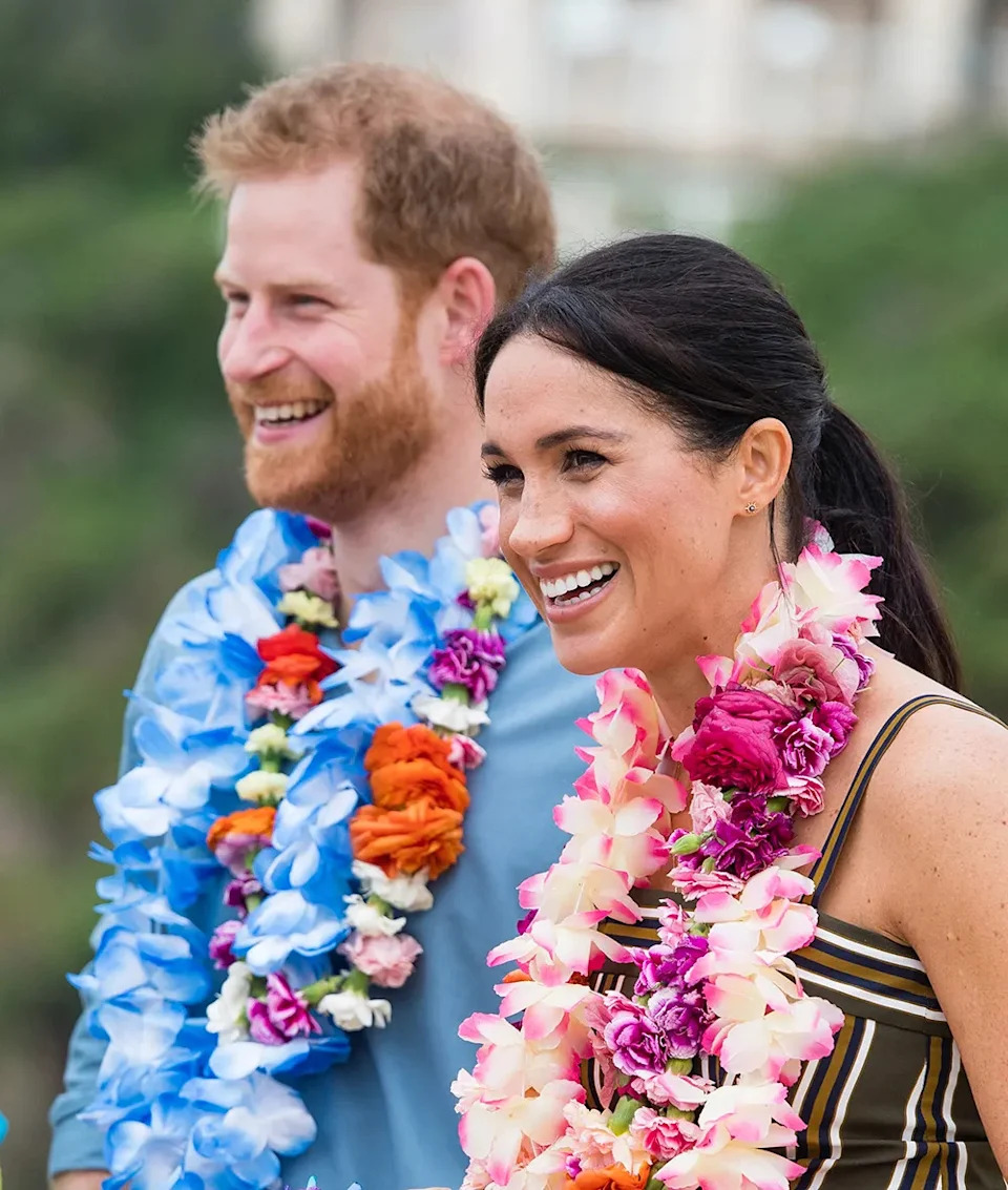 Prince Harry and Meghan Markle smiling with flower wreaths around their necks.