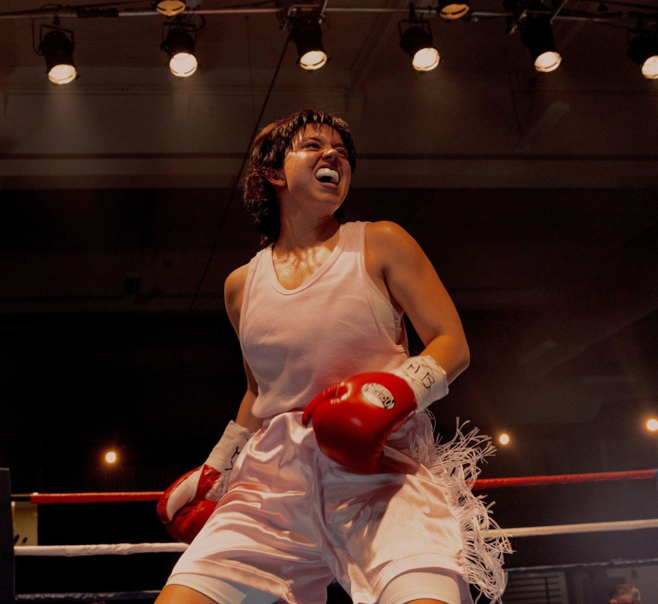A female boxer in a pink tank top and shorts celebrates in a boxing ring.