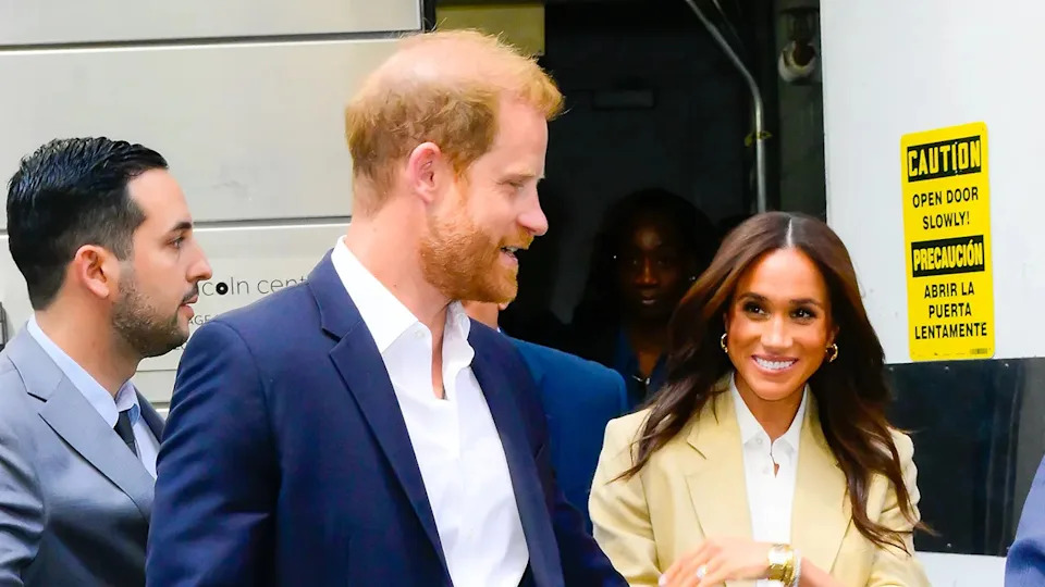 Prince Harry in a blue blazer and white shirt walking next to Meghan Markle in a beige jacket and white shirt.