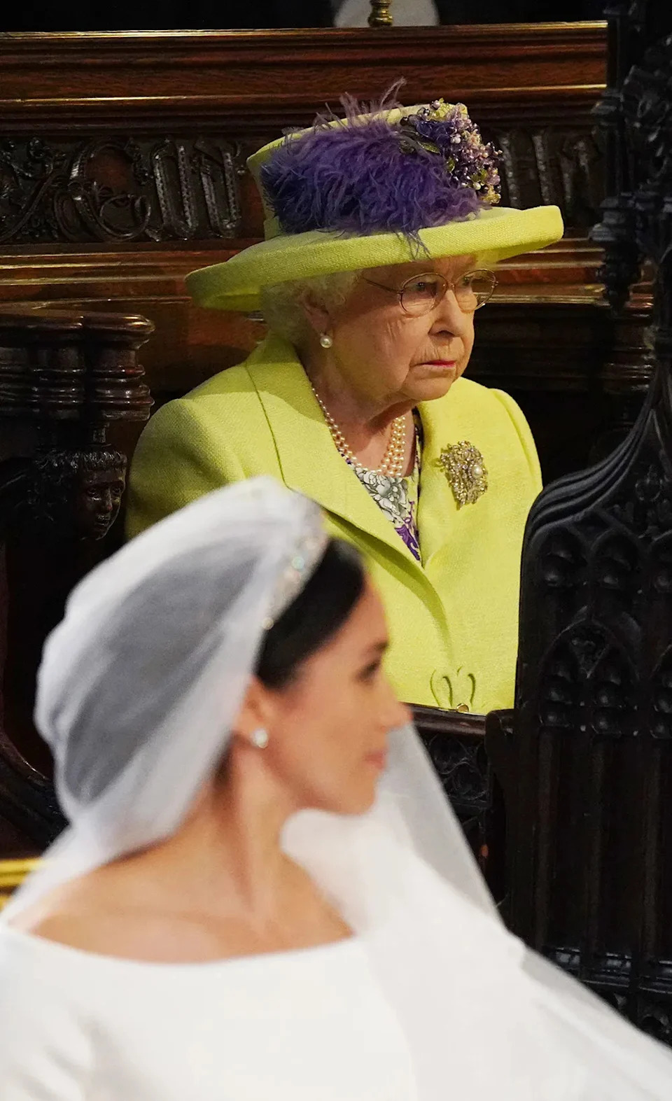 Queen Elizabeth in a bright suit looking away as Meghan Markle wears a wedding dress.