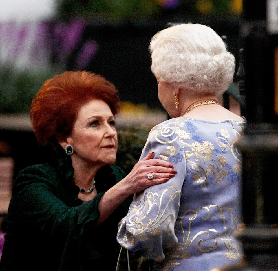 Lady Elizabeth Anson bowing down and holding on to Queen Elizabeth wearing a bright blue and ivory floral dress.