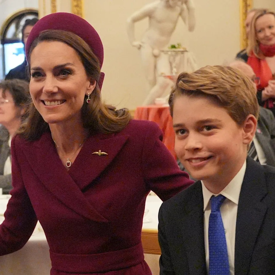 Kate Middleton wears a burgundy coat and matching hat and poses with son Prince George, who is wearing a navy suit, white shirt, and blue tie.