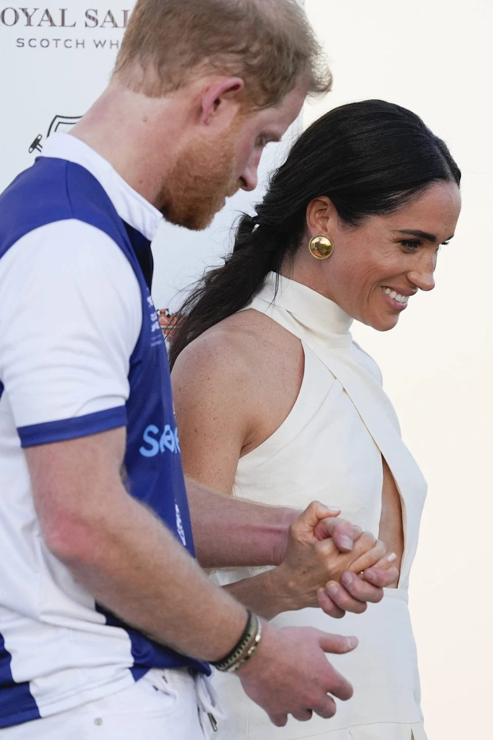 Prince Harry in a polo uniform walking alongside Meghan Markle in a cutout ivory dress as they hold hands.