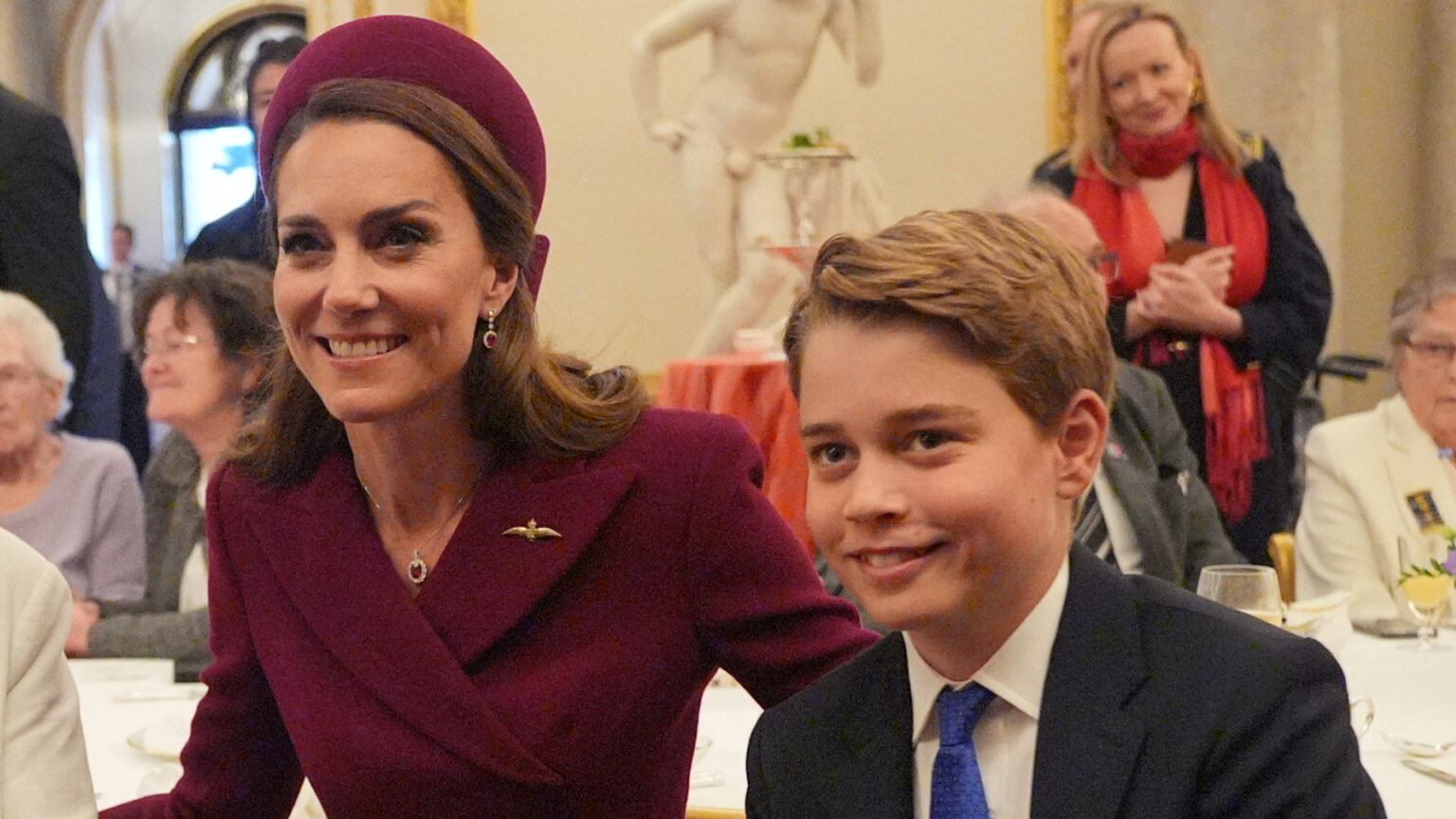 Kate Middleton wears a burgundy coat and matching hat and poses with son Prince George, who is wearing a navy suit, white shirt, and blue tie