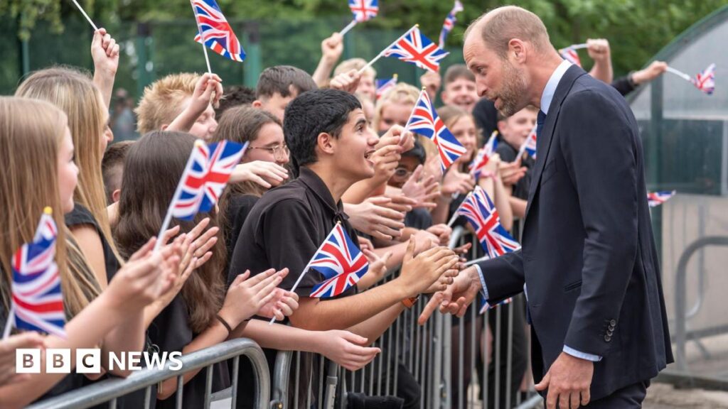 Prince William visits Sheffield school with homeless charity Prince William, dressed in a navy blue suit, is walking past a long crowd of students who are stood behind a steel fence. Many of them wave miniature Union Flags. He has stopped to speak to a male student, who has leaned over the barrier and is clapping his hands.