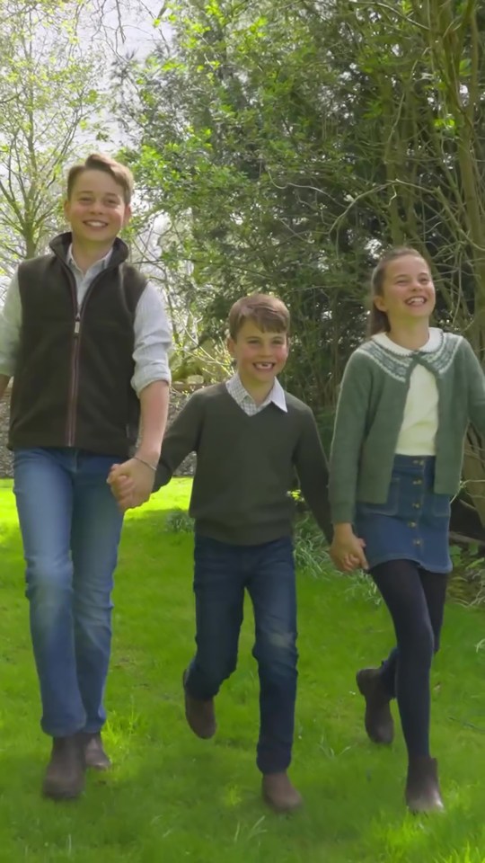 Three children holding hands and walking on a grassy lawn.