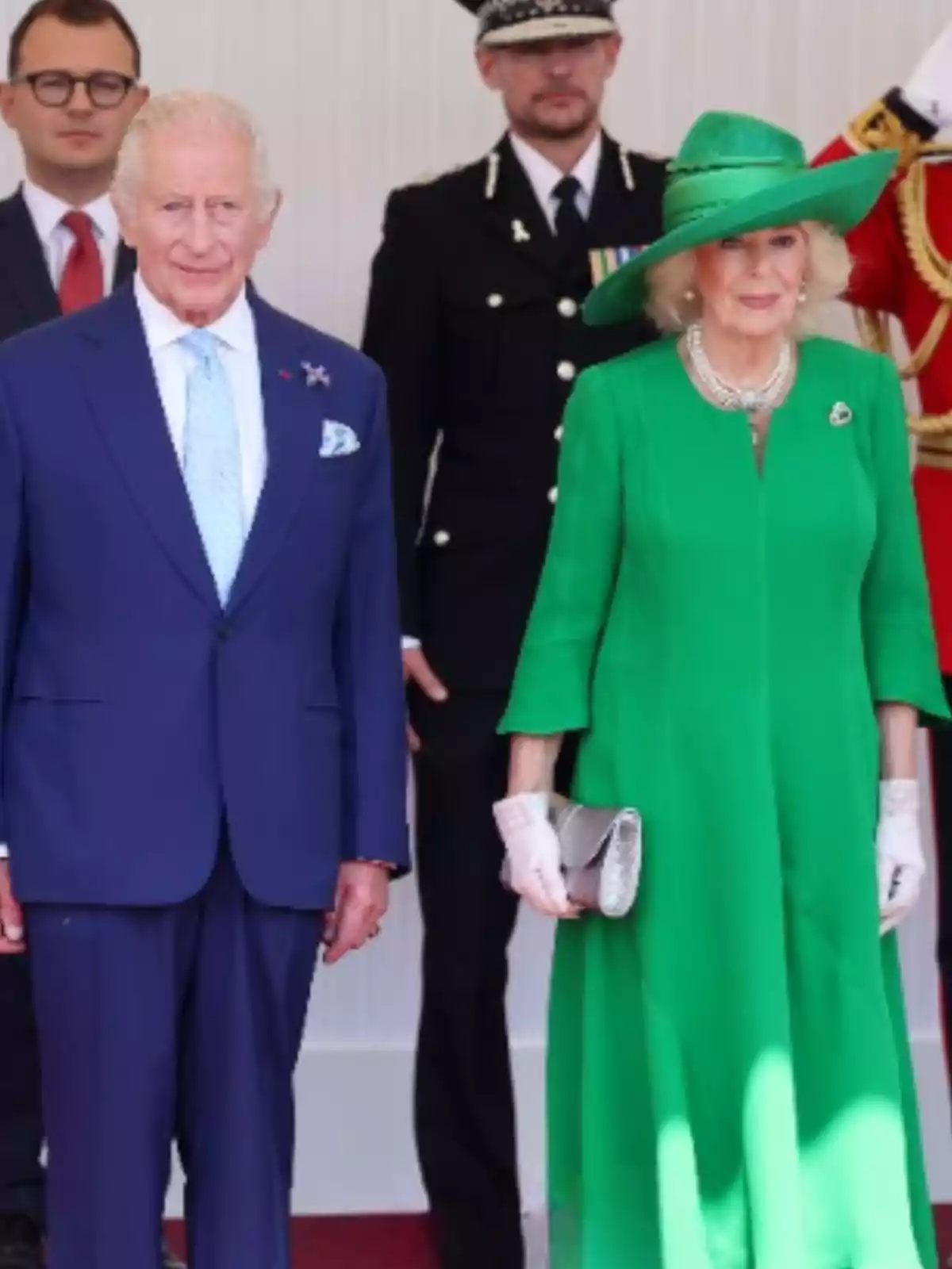 Elegantly dressed couple posing at a formal event with uniformed people in the background