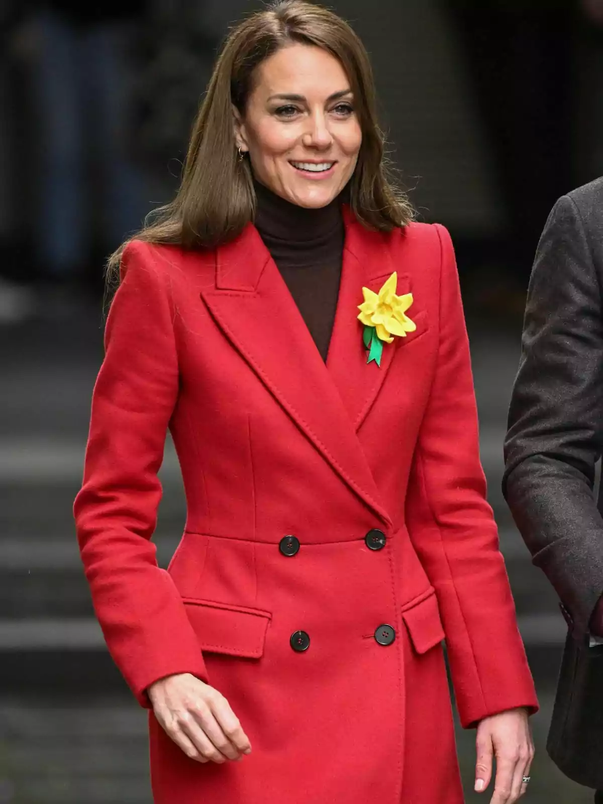 Kate Middleton in a red coat and a yellow flower brooch smiles as she walks. Kate Middleton in a red coat and a yellow flower brooch smiles as she walks.