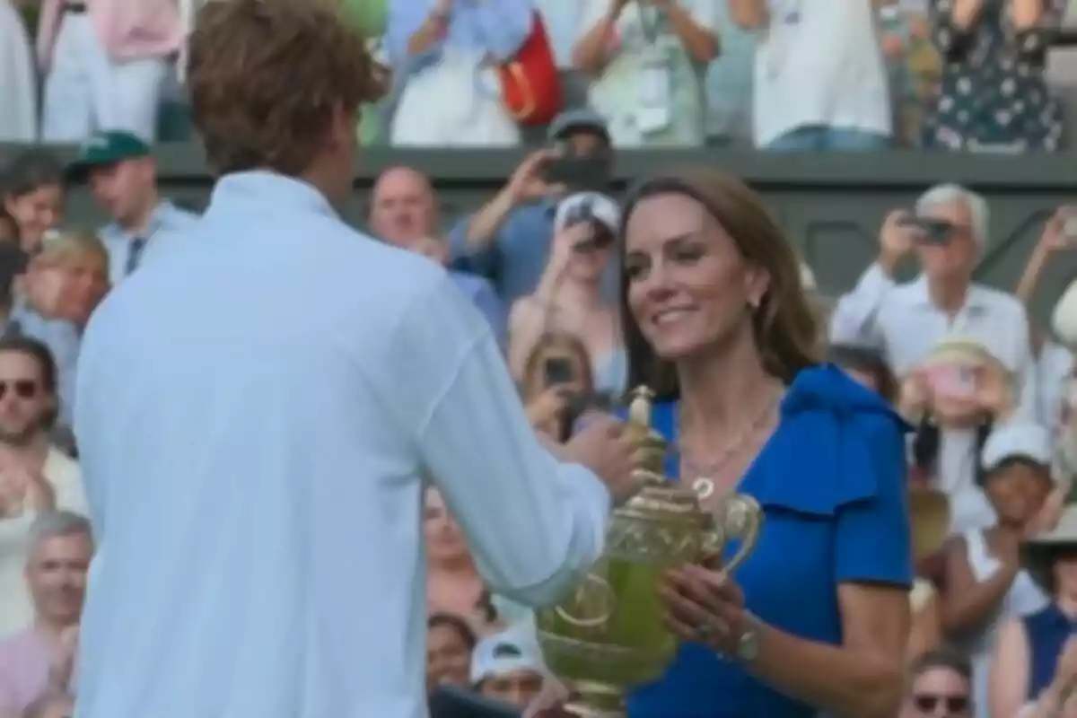 A woman in a blue dress, who is Kate Middleton, presents a golden trophy to a tennis player while the crowd watches and takes photos at Wimbledon.