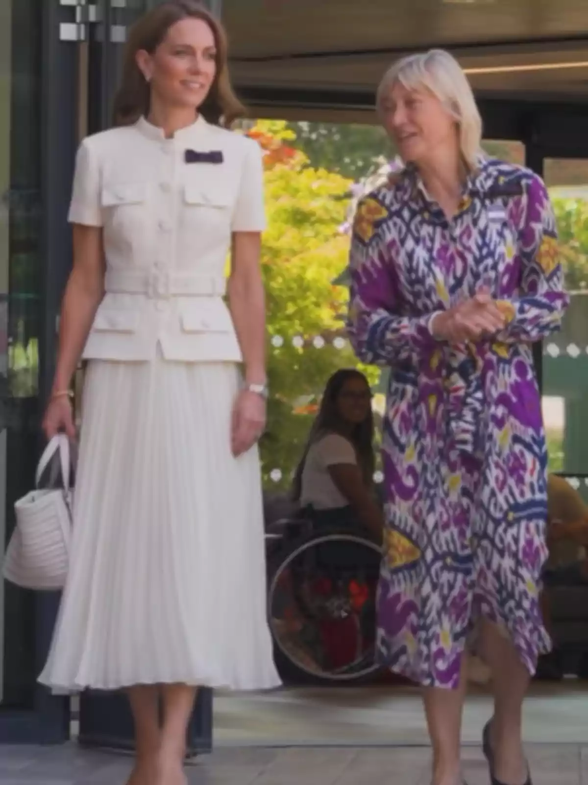 Two women walk together and talk as they leave a building—one is wearing an elegant white dress and the other a colorful dress with a geometric pattern. In the background, a person in a wheelchair can be seen.